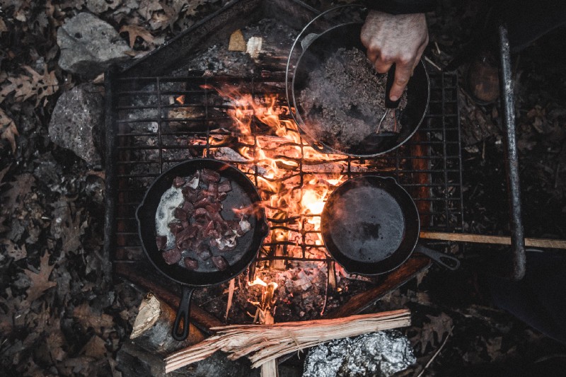 “Two cast iron pans and a pot of water over an outdoor fire-lit stove outdoors with a hand mixing the water” by Teddy Kelley on Unsplash